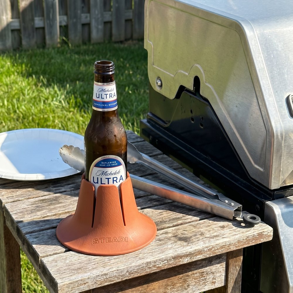 A bottle standing in a terracotta Steadi on a table outdoors on grass, a grill and fence behind.